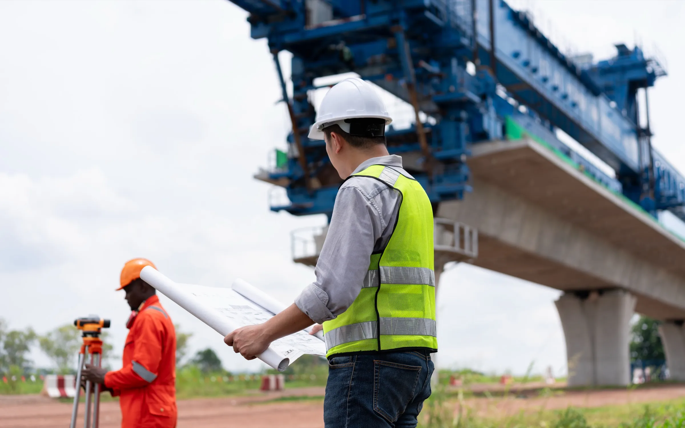 Site engineer holding rolled plans beside a surveyor at a level, in front of an elevated concrete viaduct under construction