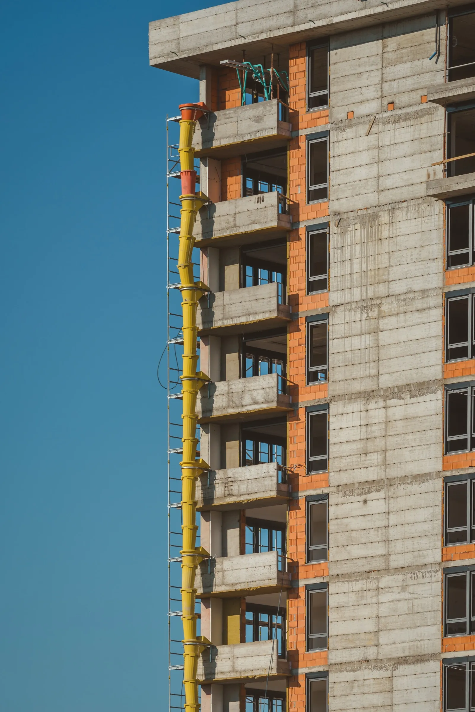 Concrete and brick residential tower under construction, a yellow debris chute running down its balconies against a blue sky
