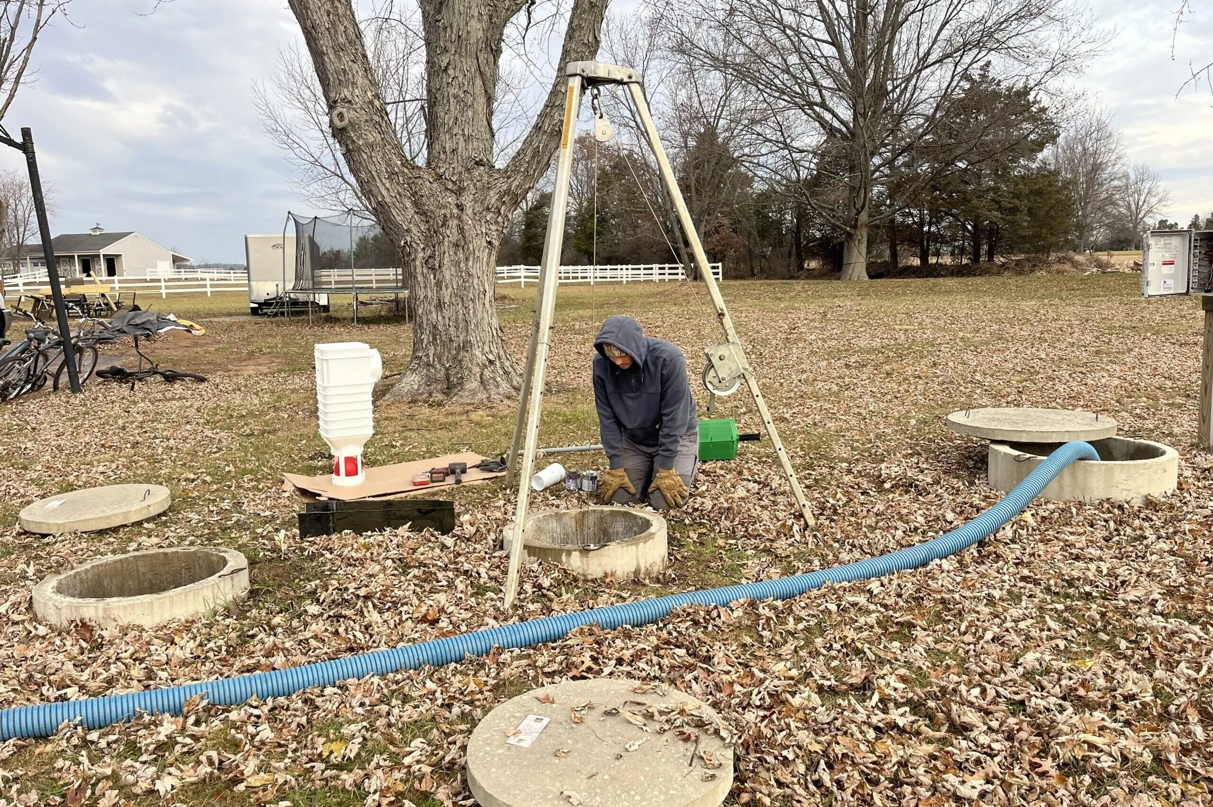Technician kneeling at a septic tank access lid in a leaf-covered yard beside a tripod hoist and a blue hose — cover image for the PreDoC website project