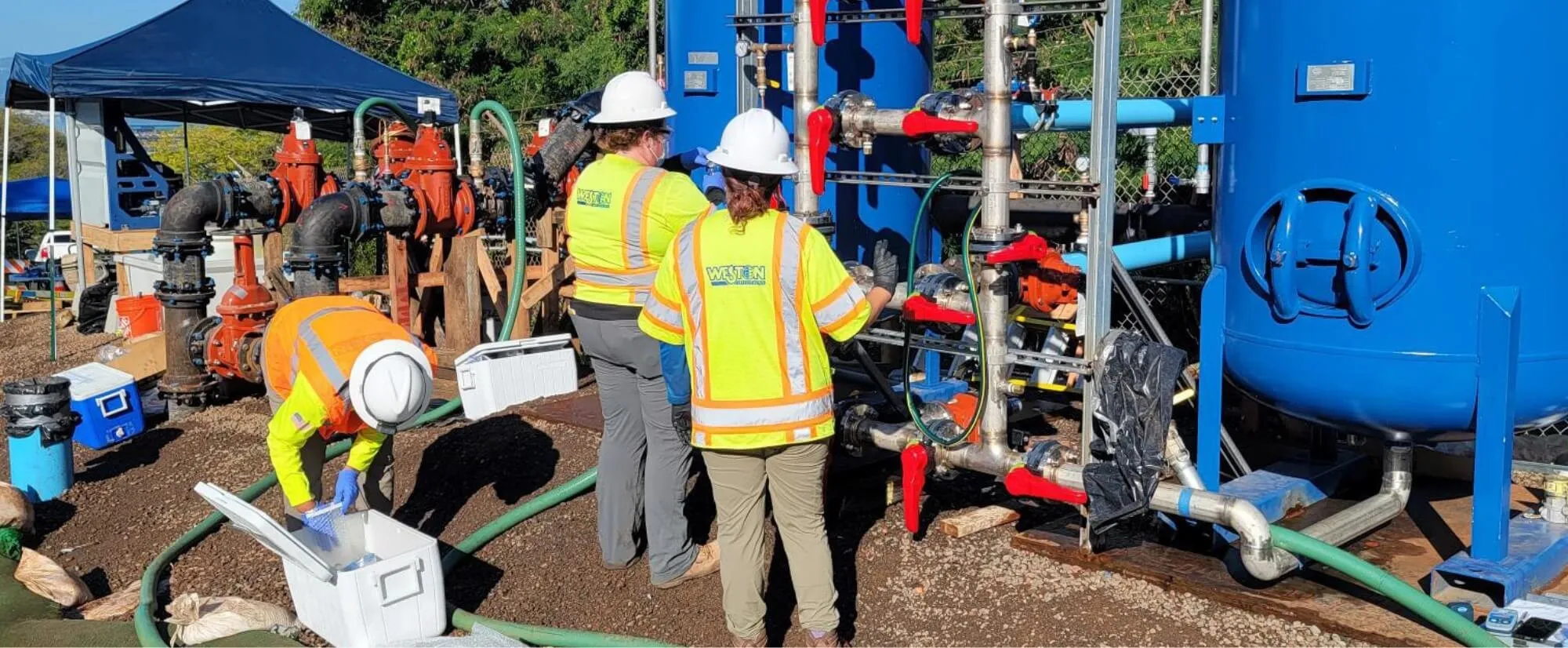 Field crew in hard hats and high-visibility vests working at blue water-treatment tanks and piping on a remediation site — cover image for the Weston Solutions website project